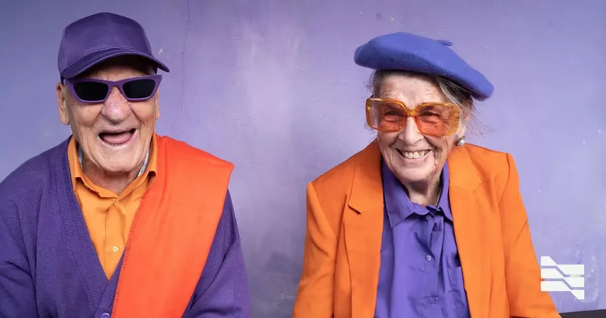 Joyful elderly couple wearing colorful outfits and trendy sunglasses against a purple wall, showcasing vibrant freelance retirement life.